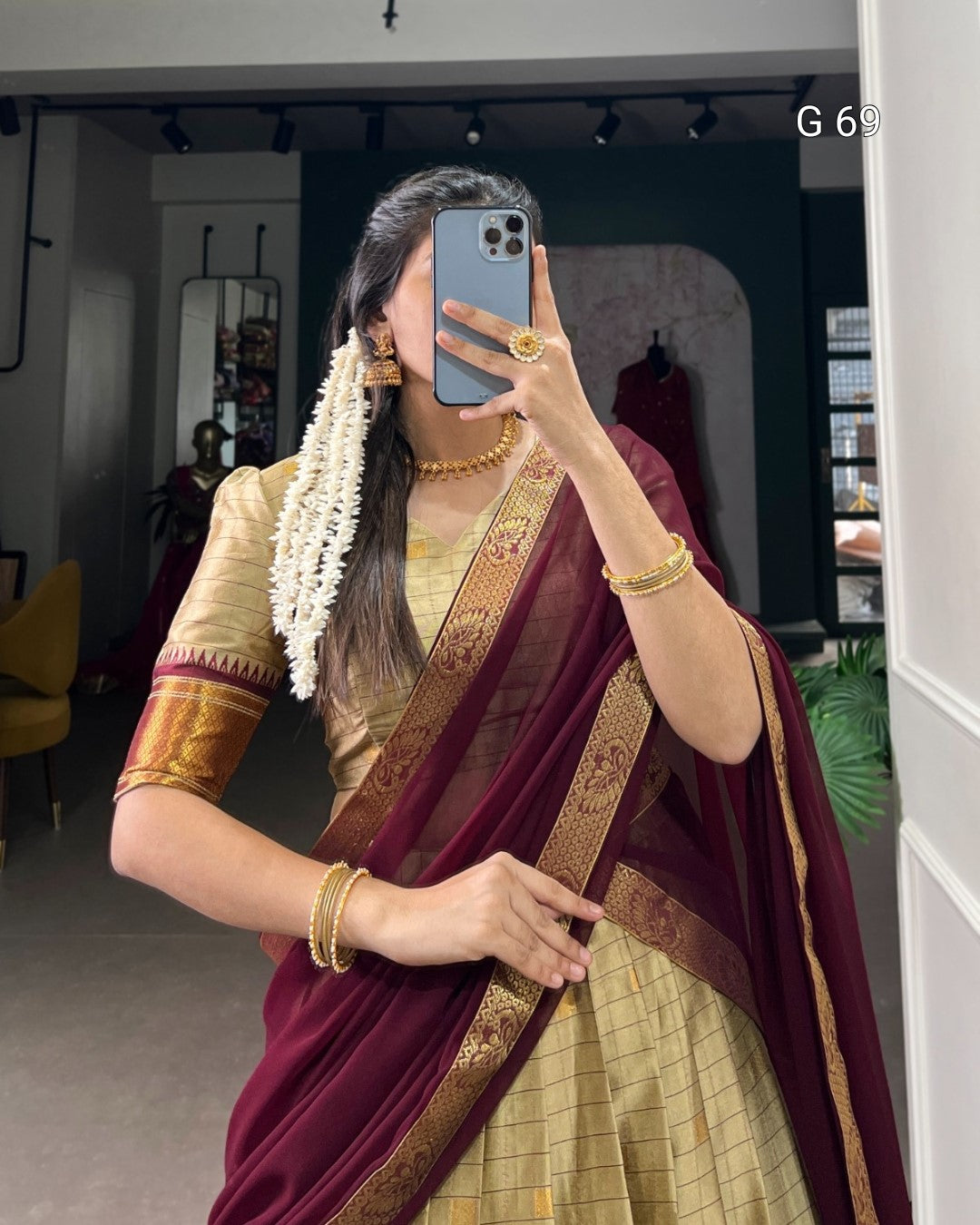 Person wearing a traditional maroon and gold saree with jewelry, taking a mirror selfie.