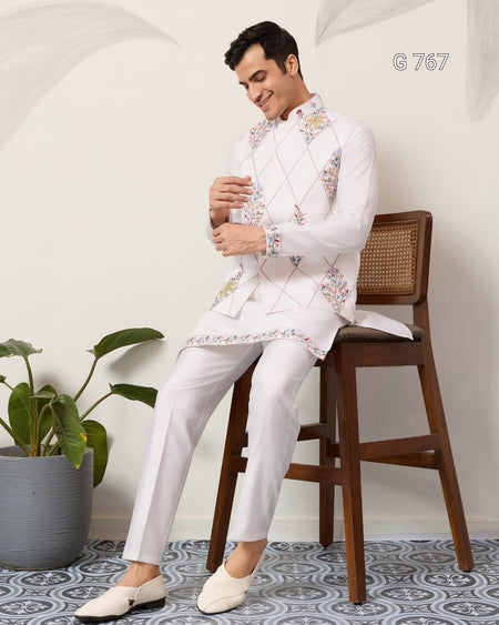 Man sitting on a wooden stool wearing a traditional embroidered vest and white pants in a minimalistic setting.