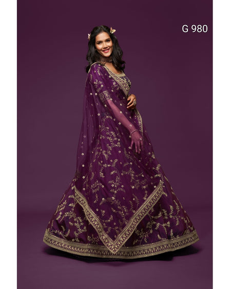 A woman modeling a traditional Indian ethnic outfit consisting of a deep purple lehenga choli with intricate gold embroidery, a matching dupatta, and a full flared skirt, set against a solid dark purple studio background. The text 'G 980' is visible in the top right corner.
