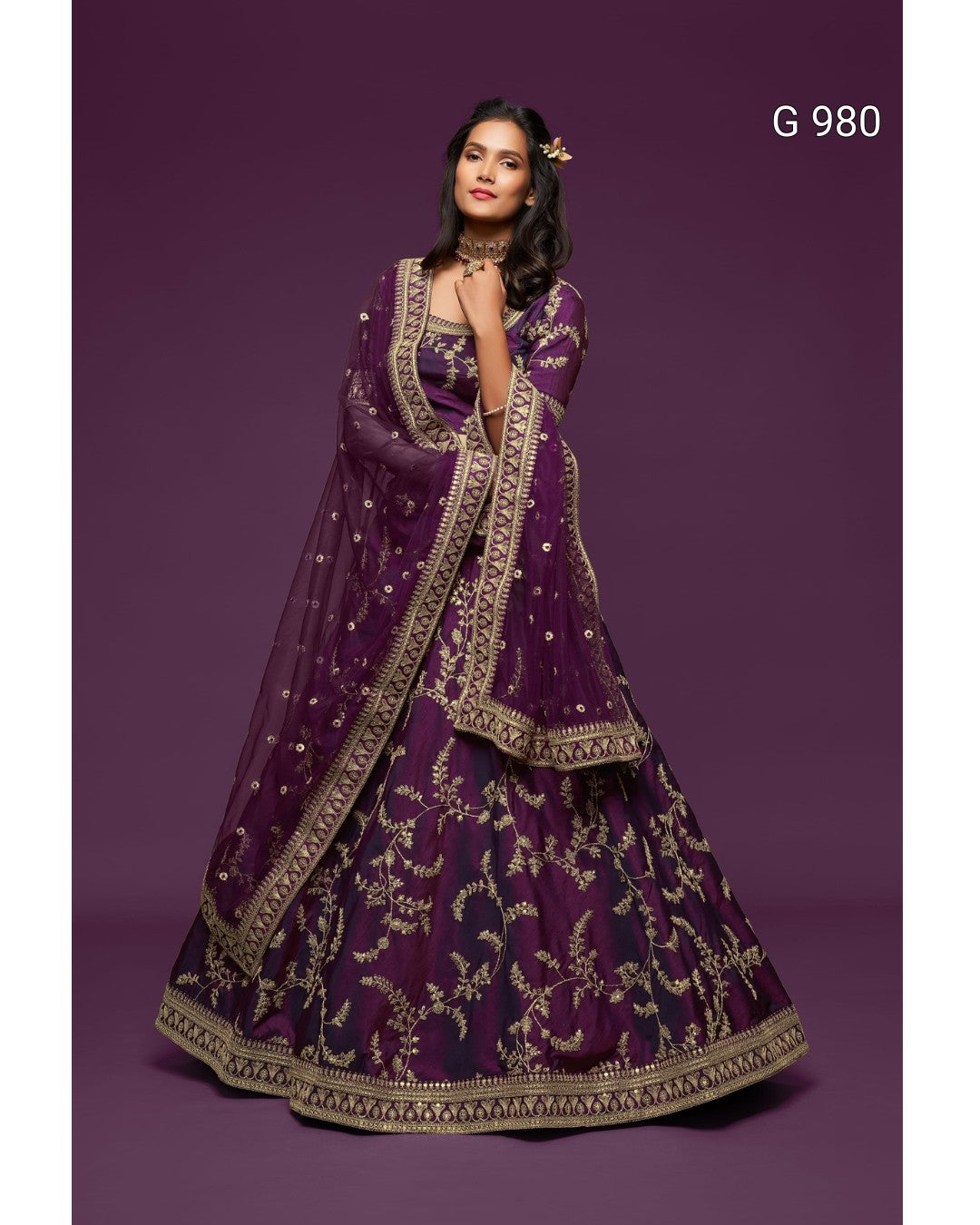A woman models a traditional Indian outfit consisting of a deep purple embroidered skirt (lehenga), matching blouse, and a sheer dupatta, all decorated with intricate gold patterns, set against a solid purple studio background.