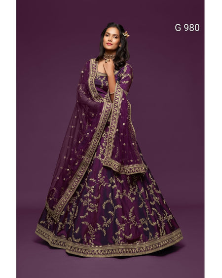 A woman models a traditional Indian outfit consisting of a deep purple embroidered skirt (lehenga), matching blouse, and a sheer dupatta, all decorated with intricate gold patterns, set against a solid purple studio background.
