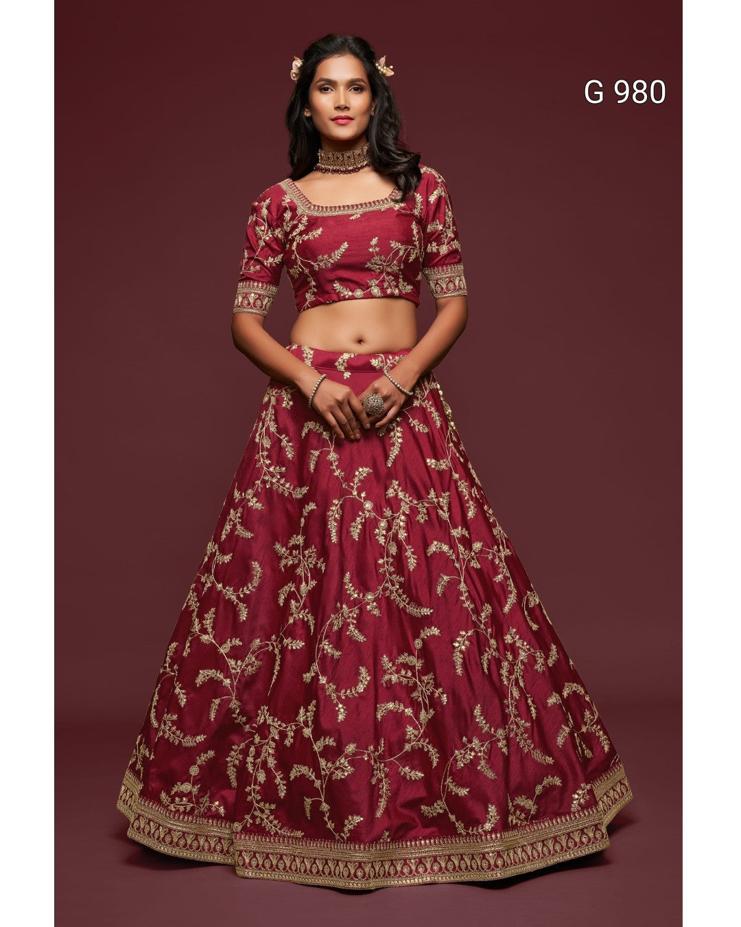 A woman modeling a traditional Indian lehenga choli set in deep maroon with intricate gold embroidery, set against a solid maroon studio background. The text 'G 980' is visible in the top right corner.