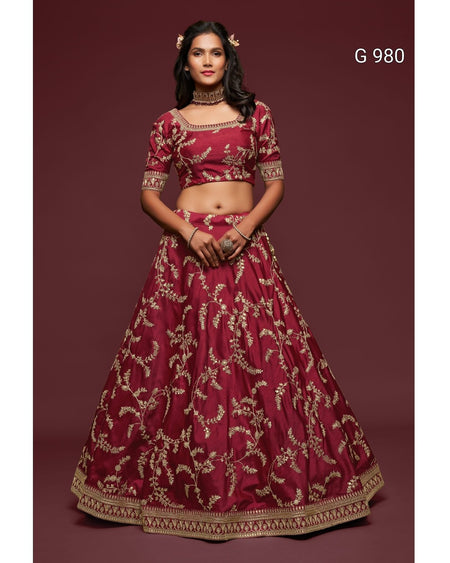 A woman modeling a traditional Indian lehenga choli set in deep maroon with intricate gold embroidery, set against a solid maroon studio background. The text 'G 980' is visible in the top right corner.