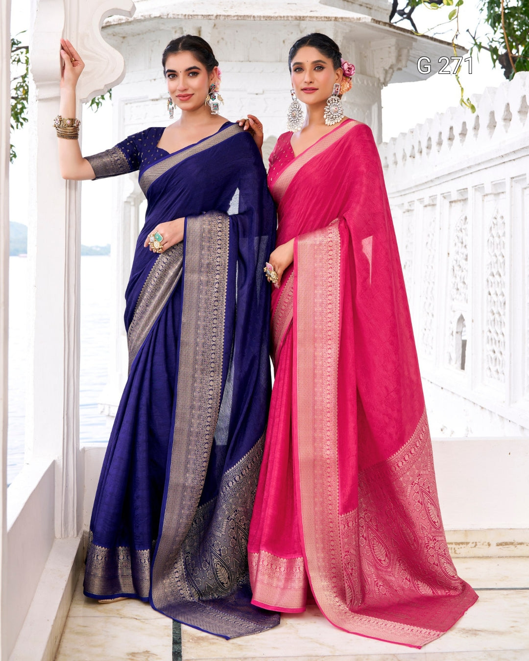 Two women modeling traditional Indian sarees in a white architectural setting. One saree is deep navy blue with a silver border, and the other is bright pink with a gold border.