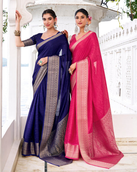 Two women modeling traditional Indian sarees in a white architectural setting. One saree is deep navy blue with a silver border, and the other is bright pink with a gold border.