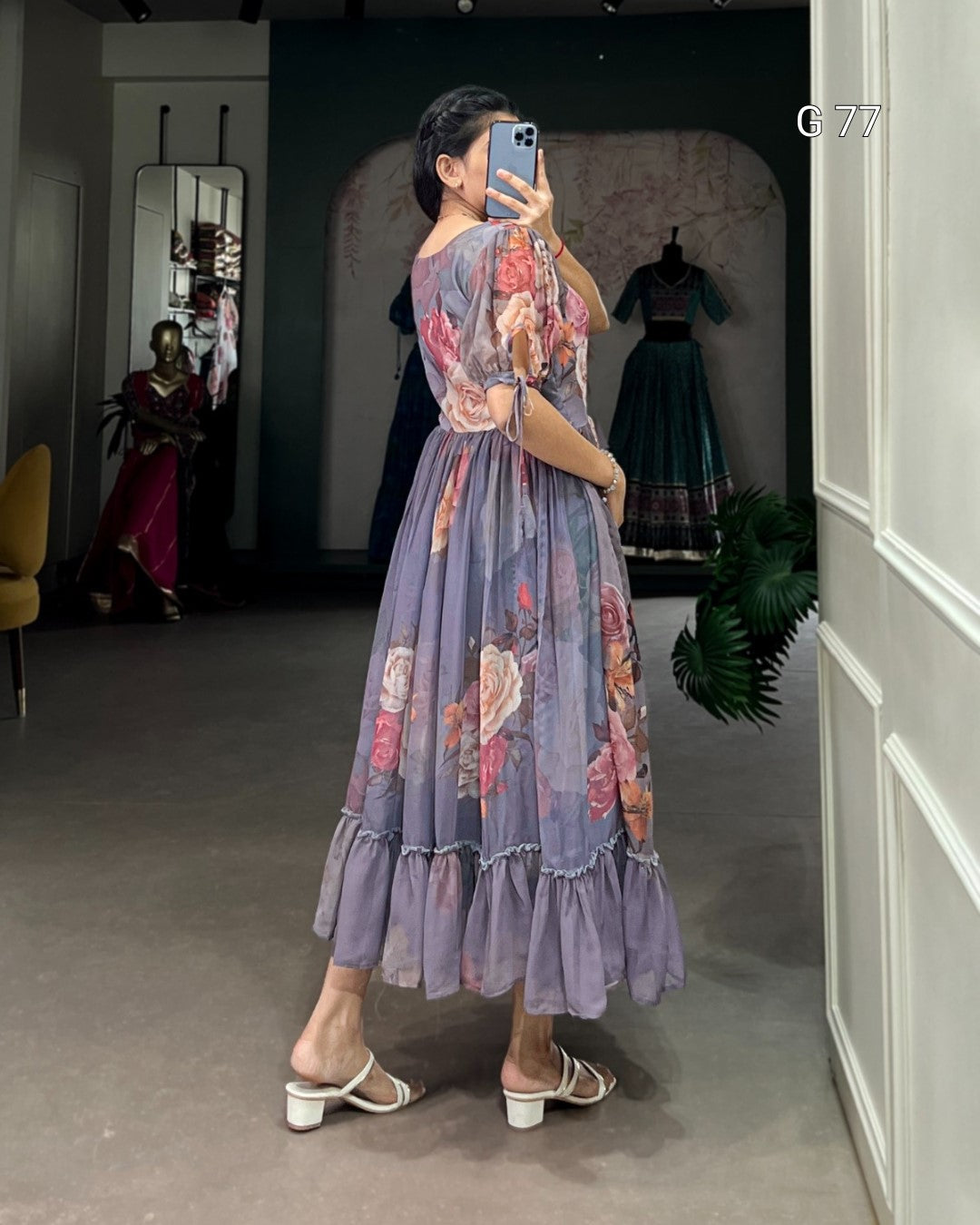 Woman taking a mirror selfie wearing a floral dress in a room with a plant and another person in the background.
