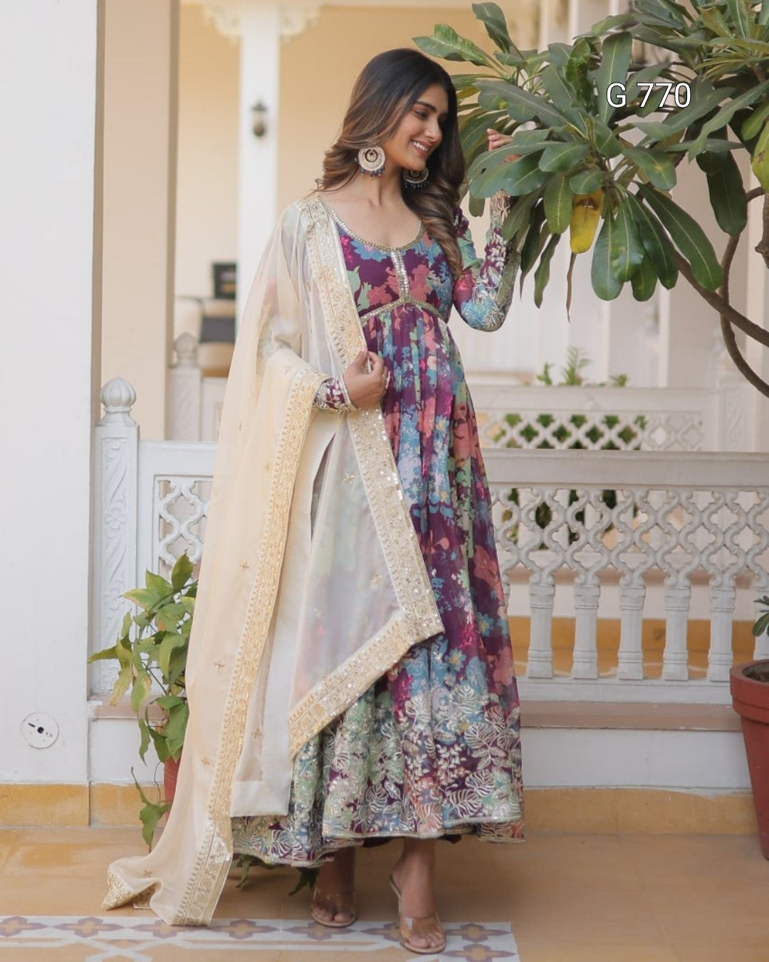 Woman in a floral dress with a beige dupatta standing indoors with plants around.