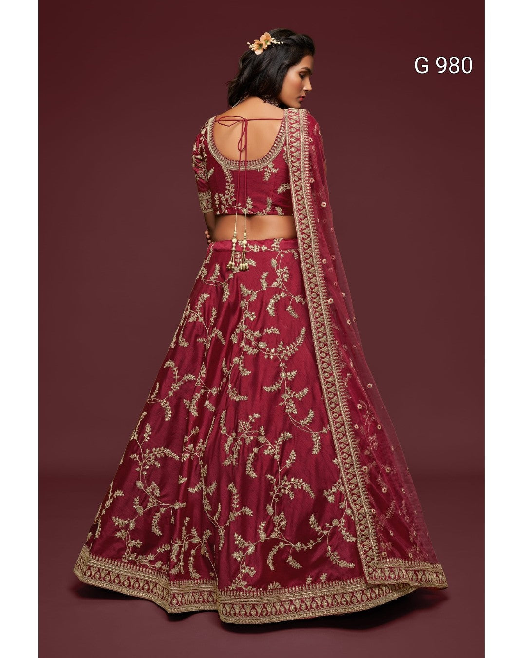 A woman modeling a deep maroon lehenga choli set with intricate gold embroidery, shown from the back against a solid maroon background. The set includes a flared skirt, a cropped embroidered blouse, and a matching dupatta.