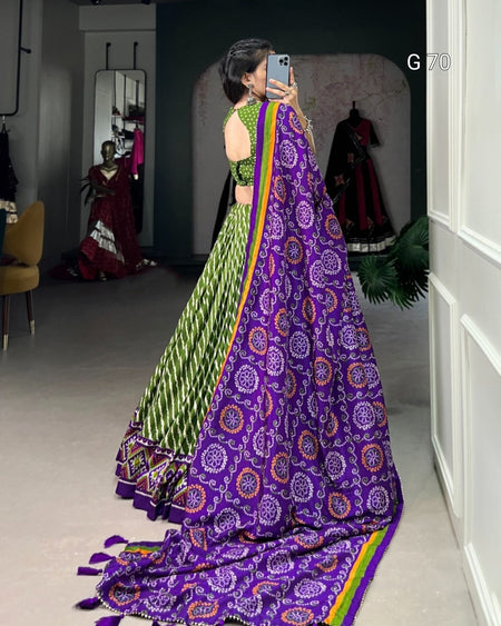 Person wearing a green and purple traditional outfit with a colorful border, standing in a room.