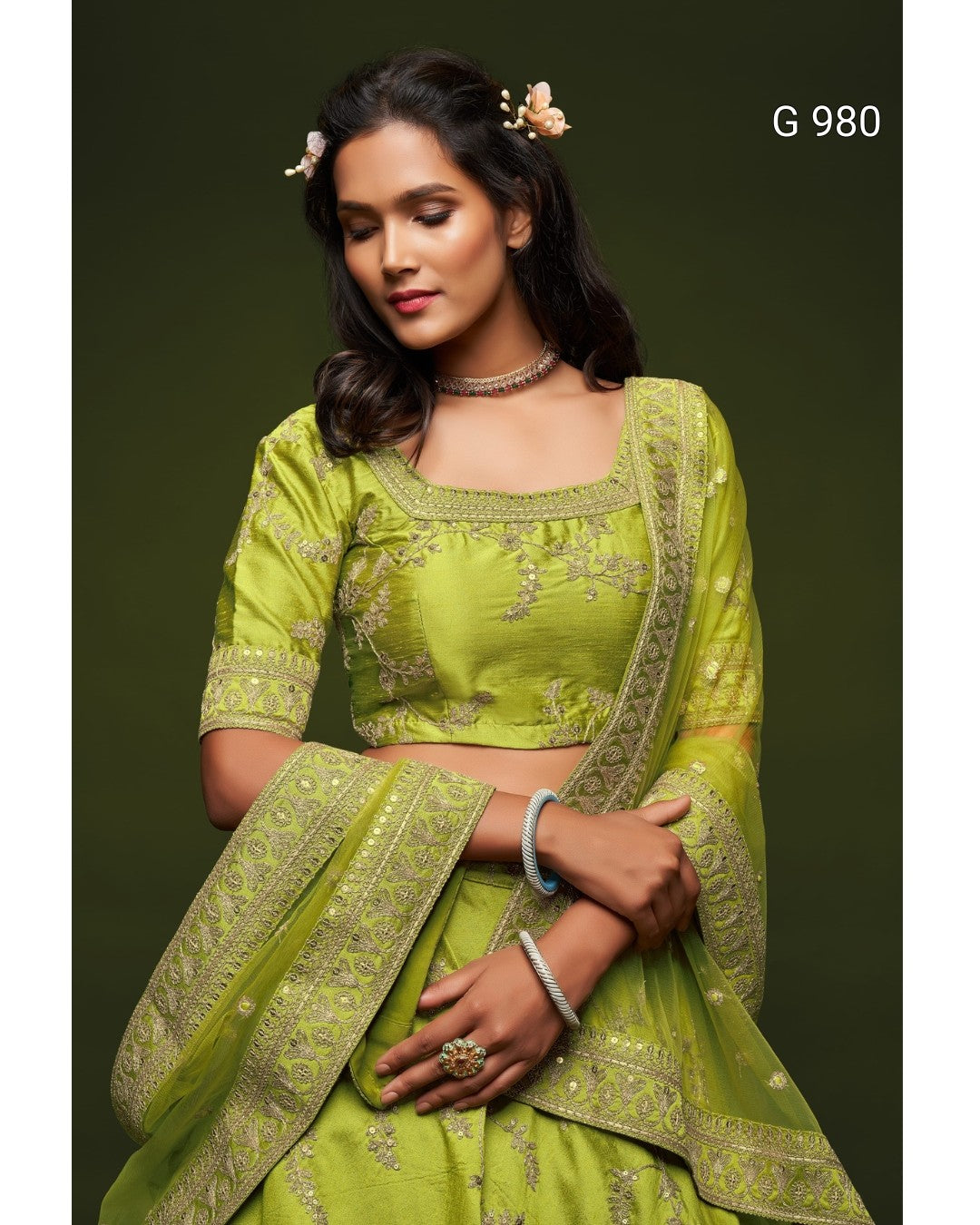 A woman modeling a lime green traditional Indian outfit with matching embroidered dupatta against a dark olive green background. The outfit features a cropped blouse with short sleeves, a square neckline, and detailed gold embroidery.
