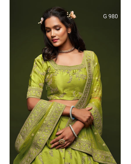 A woman modeling a lime green traditional Indian outfit with matching embroidered dupatta against a dark olive green background. The outfit features a cropped blouse with short sleeves, a square neckline, and detailed gold embroidery.