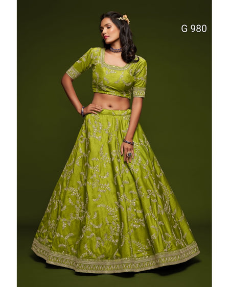 A woman modeling a lime green traditional Indian lehenga choli set with gold floral embroidery, set against a solid dark olive green background. The text 'G 980' is visible in the top right corner.
