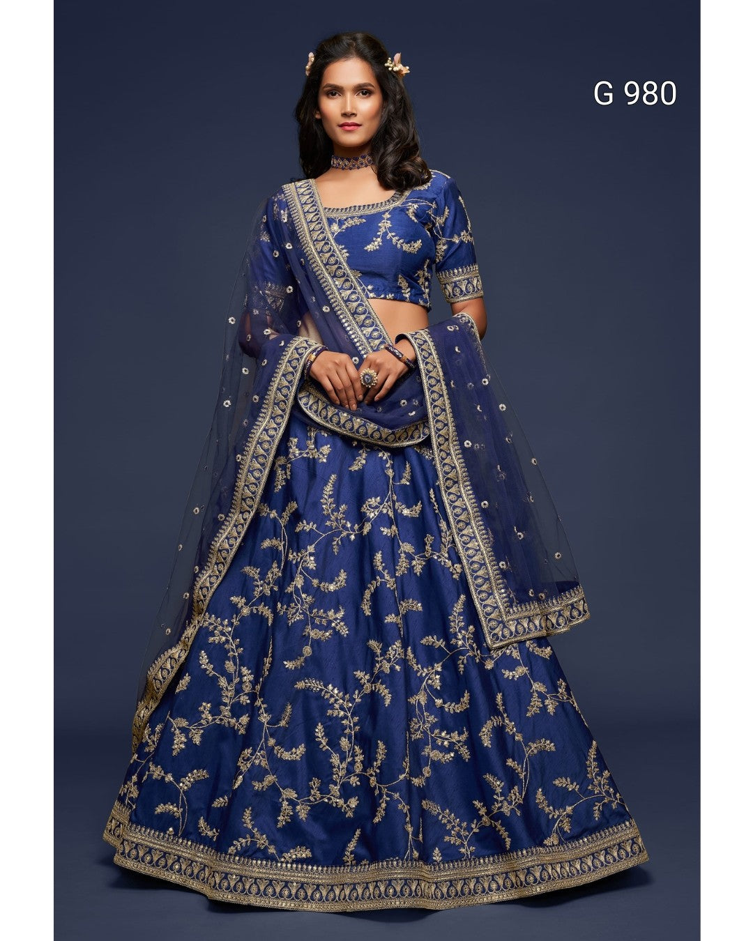 A woman modeling a royal blue lehenga choli with intricate gold embroidery, set against a dark blue studio background. The text 'G 980' is visible in the top right corner.
