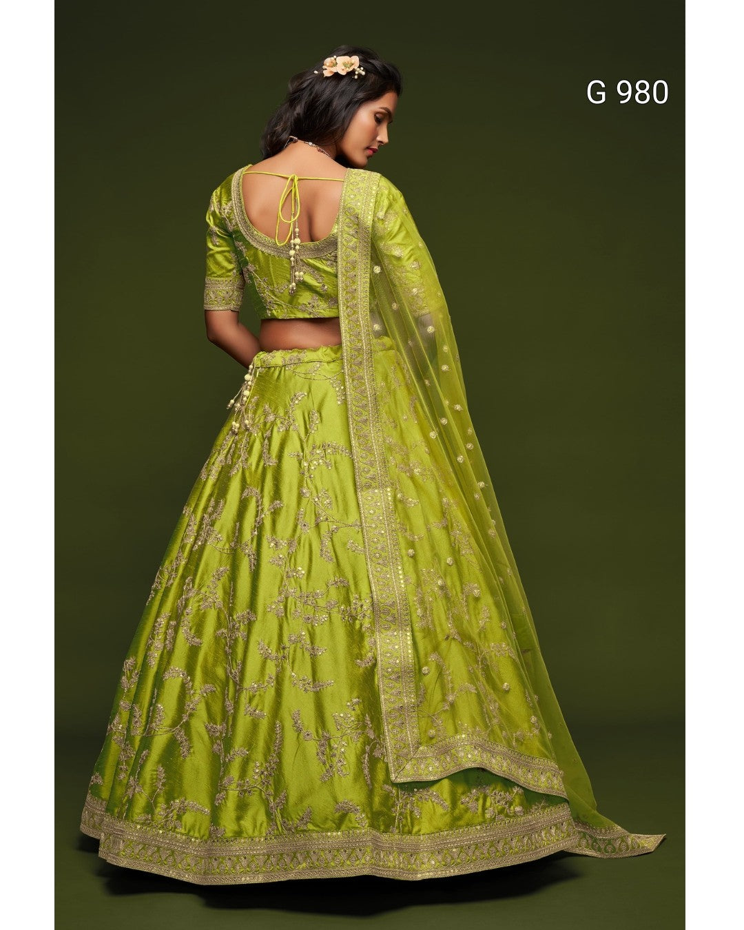 A woman modeling a bright lime green embroidered lehenga choli set with a matching sheer dupatta, shown from the back against a dark olive green studio background. The set features intricate gold embroidery and has the text 'G 980' in the top right corner.