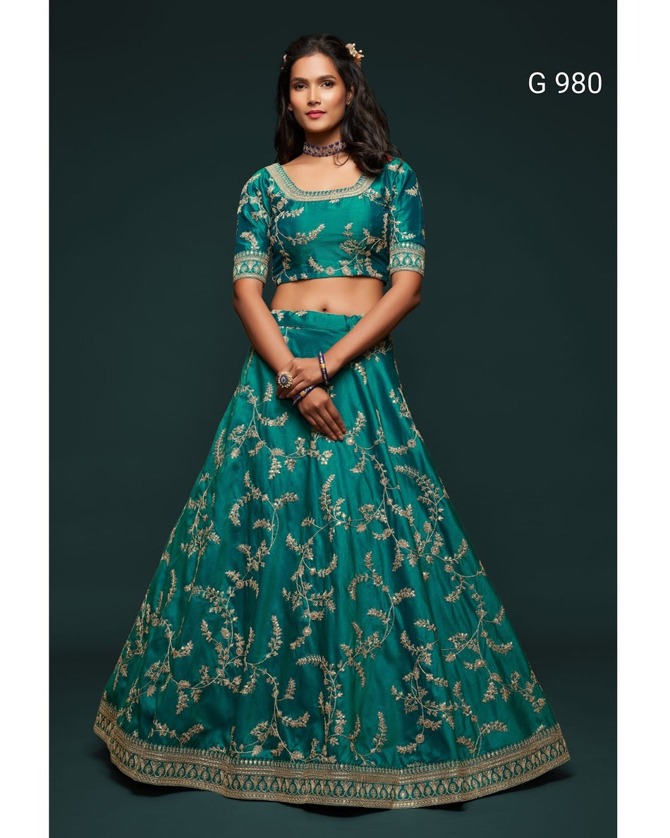 A woman modeling a teal green lehenga choli set with intricate gold floral embroidery, set against a dark green background. The text 'G 980' is visible in the top right corner.