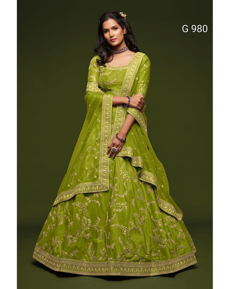A woman models a lime green traditional Indian lehenga choli set with gold embroidery against a dark olive green studio background. The set includes a flared skirt (lehenga), a matching blouse (choli), and a dupatta.