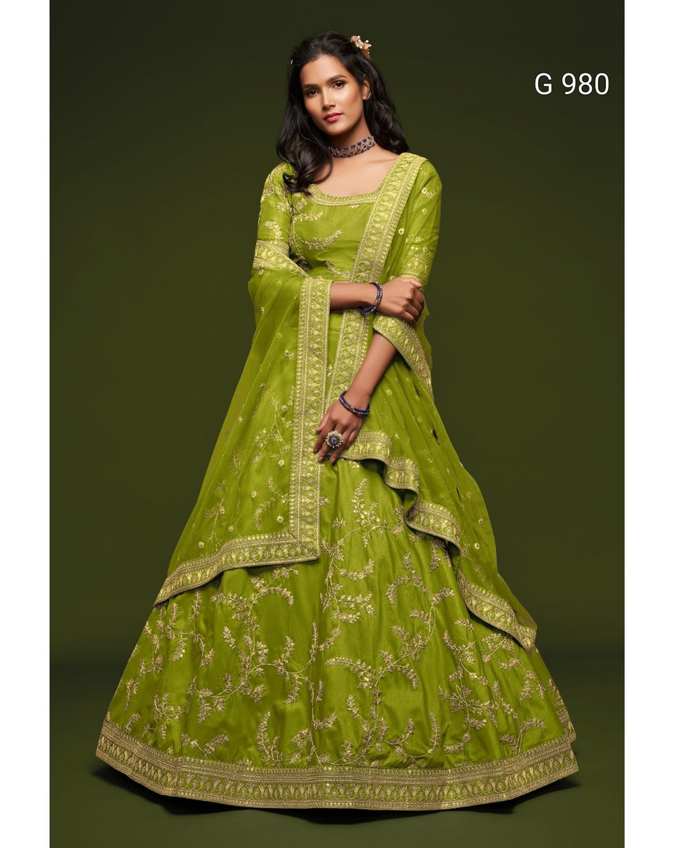 A woman models a lime green traditional Indian lehenga choli set with gold embroidery against a dark olive green studio background. The set includes a flared skirt (lehenga), a matching blouse (choli), and a dupatta.