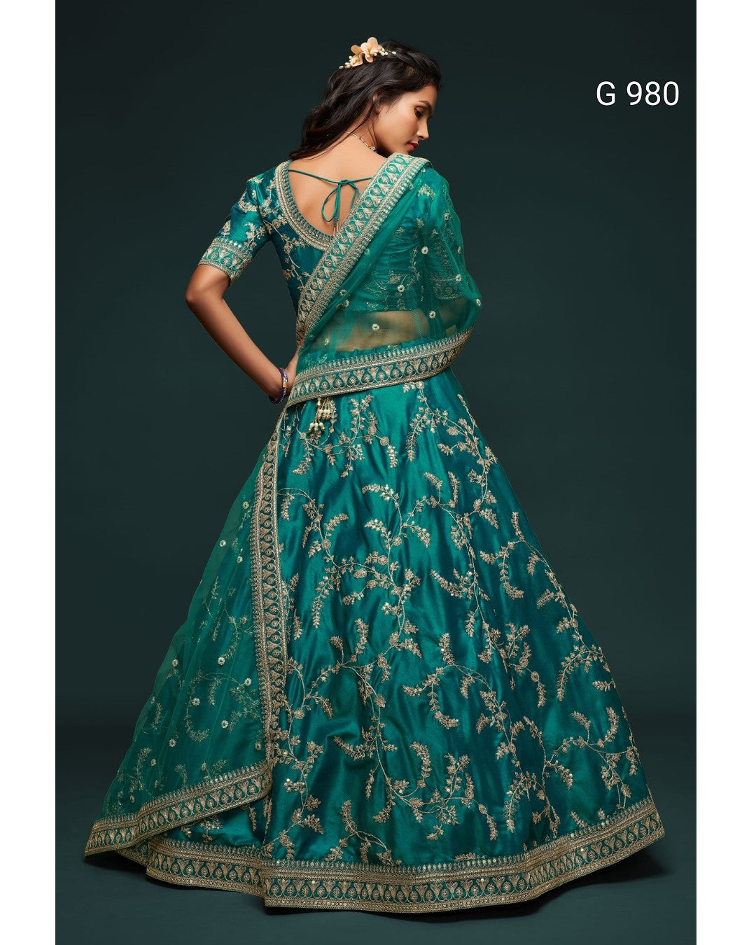 A woman modeling a teal green lehenga choli set with intricate gold embroidery, shown from the back against a dark green background. The text 'G 980' is visible in the top right corner.