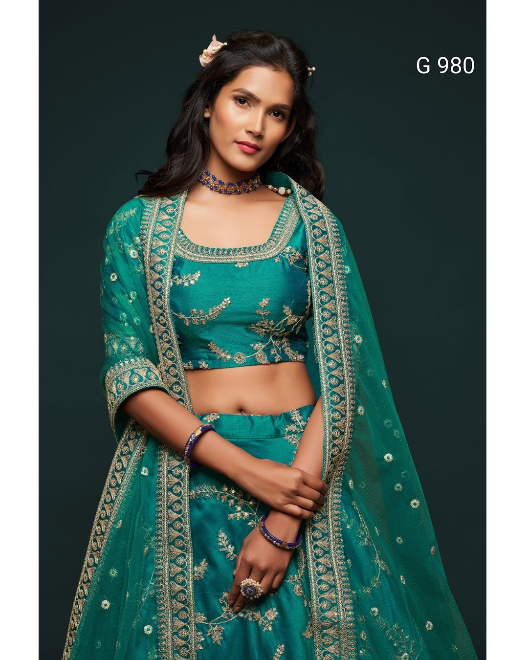 A woman modeling a teal green embroidered Indian ethnic outfit with a matching dupatta, set against a dark green background. The outfit features intricate gold floral embroidery and is labeled with the code 'G 980' in the top right corner.