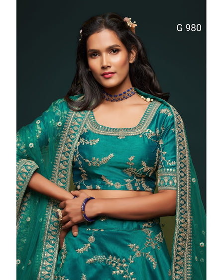 A woman modeling a teal green embroidered traditional outfit with a matching sheer dupatta, featuring gold floral embroidery and mirror work, set against a dark background with the text 'G 980' in the top right.
