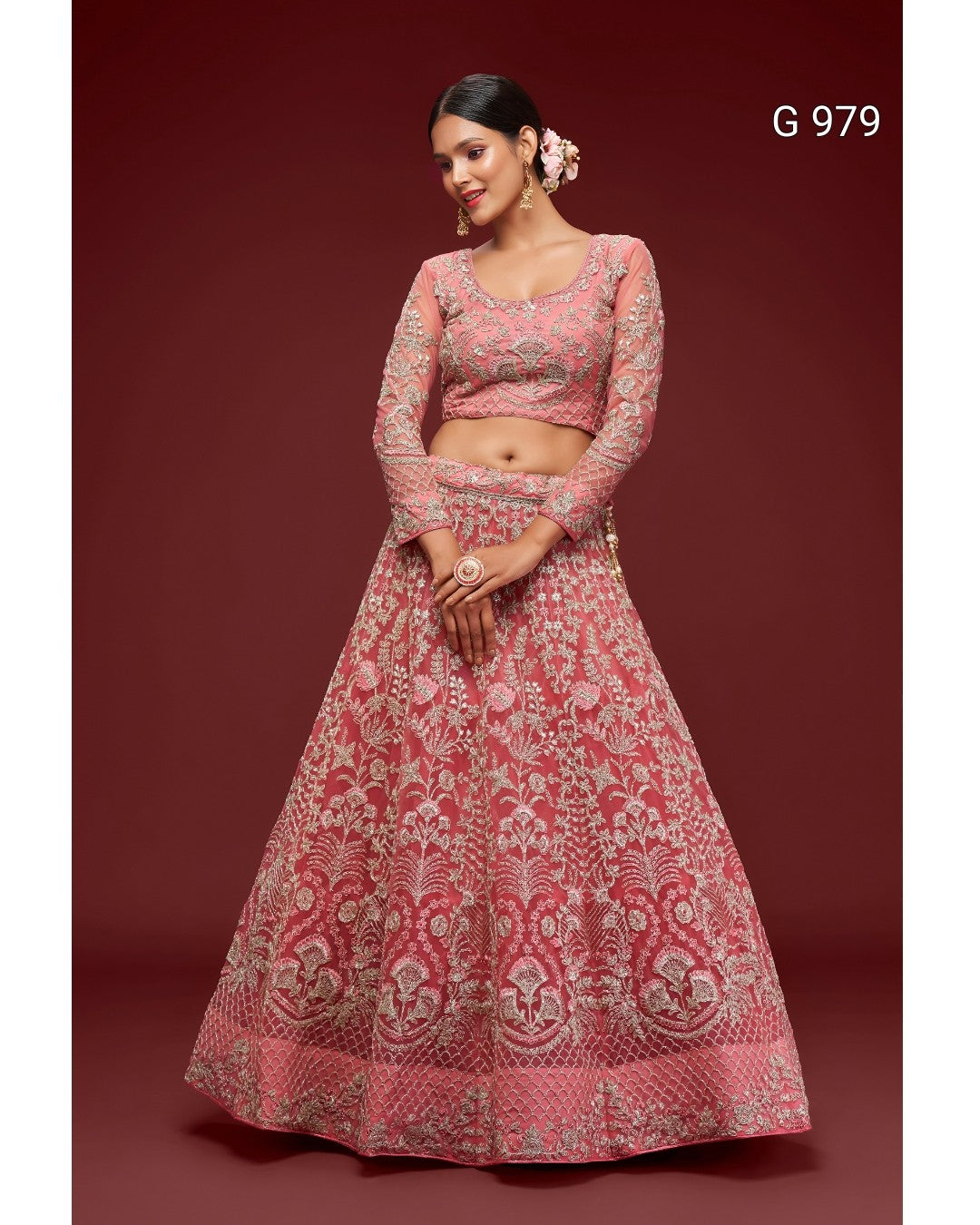 A woman models a pink and gold heavily embroidered Indian ethnic outfit, consisting of a long-sleeved crop top and a matching flared skirt, set against a solid dark red background.