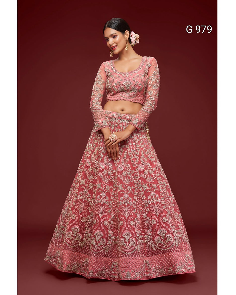 A woman models a pink and gold heavily embroidered Indian ethnic outfit, consisting of a long-sleeved crop top and a matching flared skirt, set against a solid dark red background.