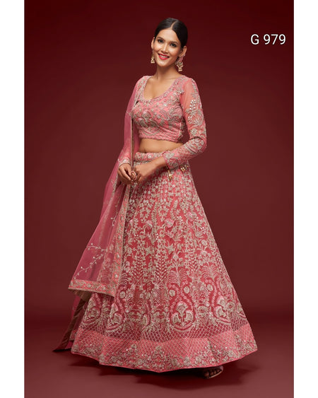 A woman models a pink and gold heavily embroidered traditional Indian lehenga choli set against a solid dark red-brown studio background. The set features a pink embroidered blouse with long sleeves, a matching flared skirt, and a sheer dupatta.