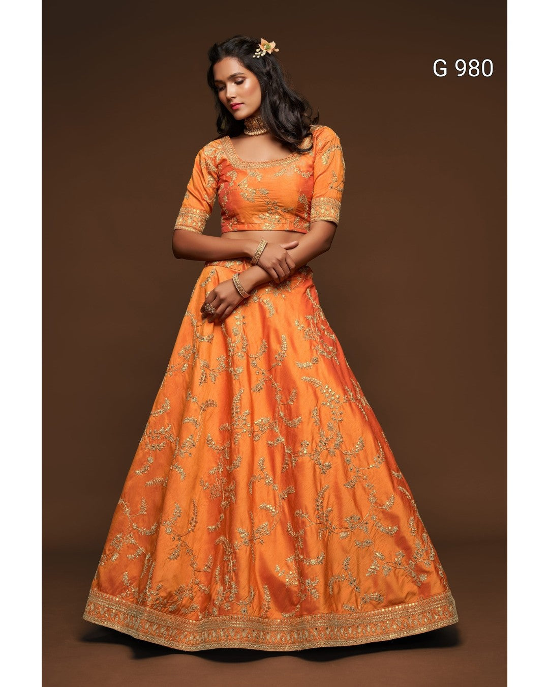 An orange traditional Indian lehenga choli set with gold floral embroidery, modeled against a solid brown studio background. The text 'G 980' is visible in the top right corner.