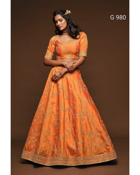 An orange traditional Indian lehenga choli set with gold floral embroidery, modeled against a solid brown studio background. The text 'G 980' is visible in the top right corner.