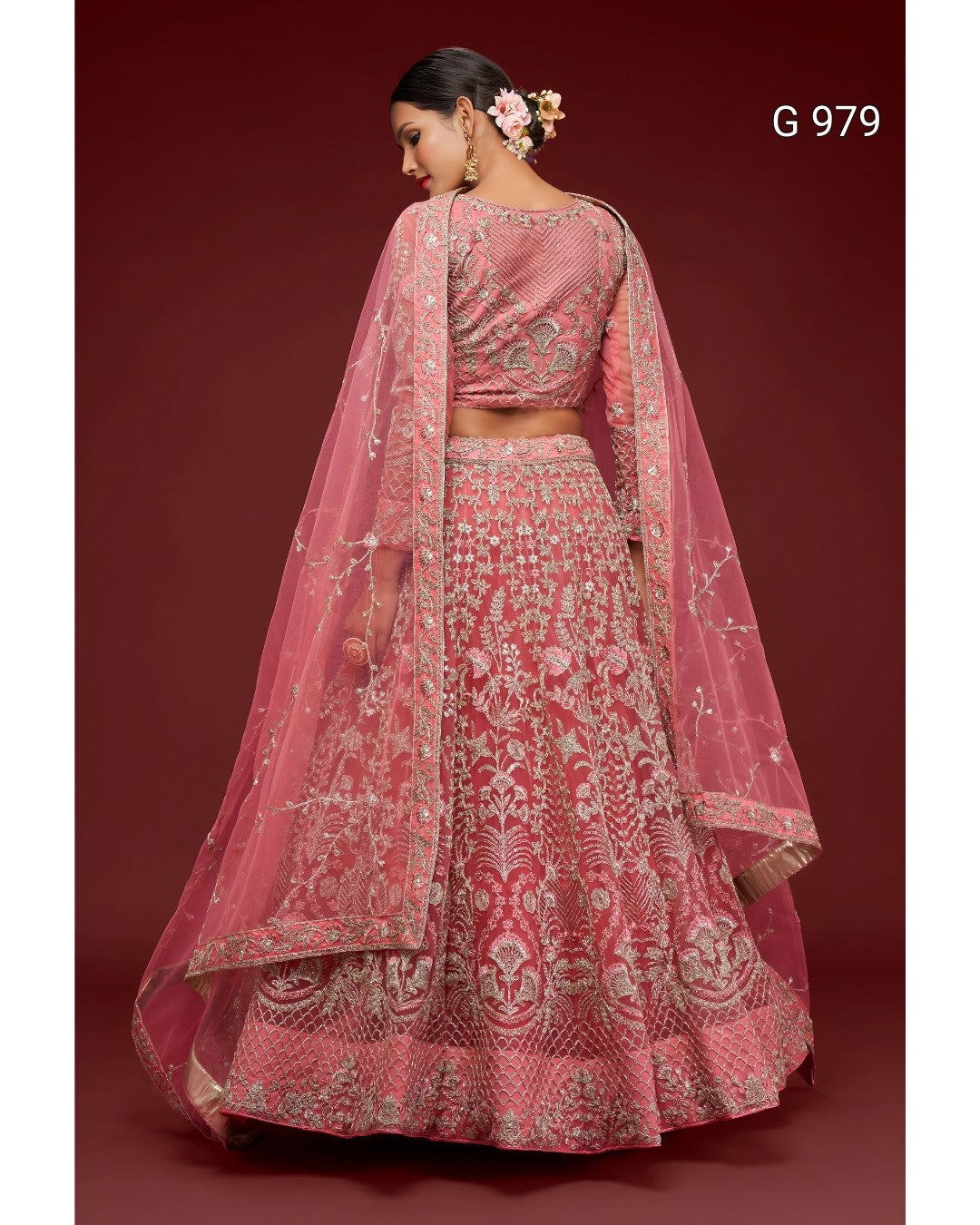A pink heavily-embroidered Indian ethnic lehenga choli set with a matching dupatta, shown on a model from the back against a dark red background. The set features intricate silver and gold embroidery with floral motifs.