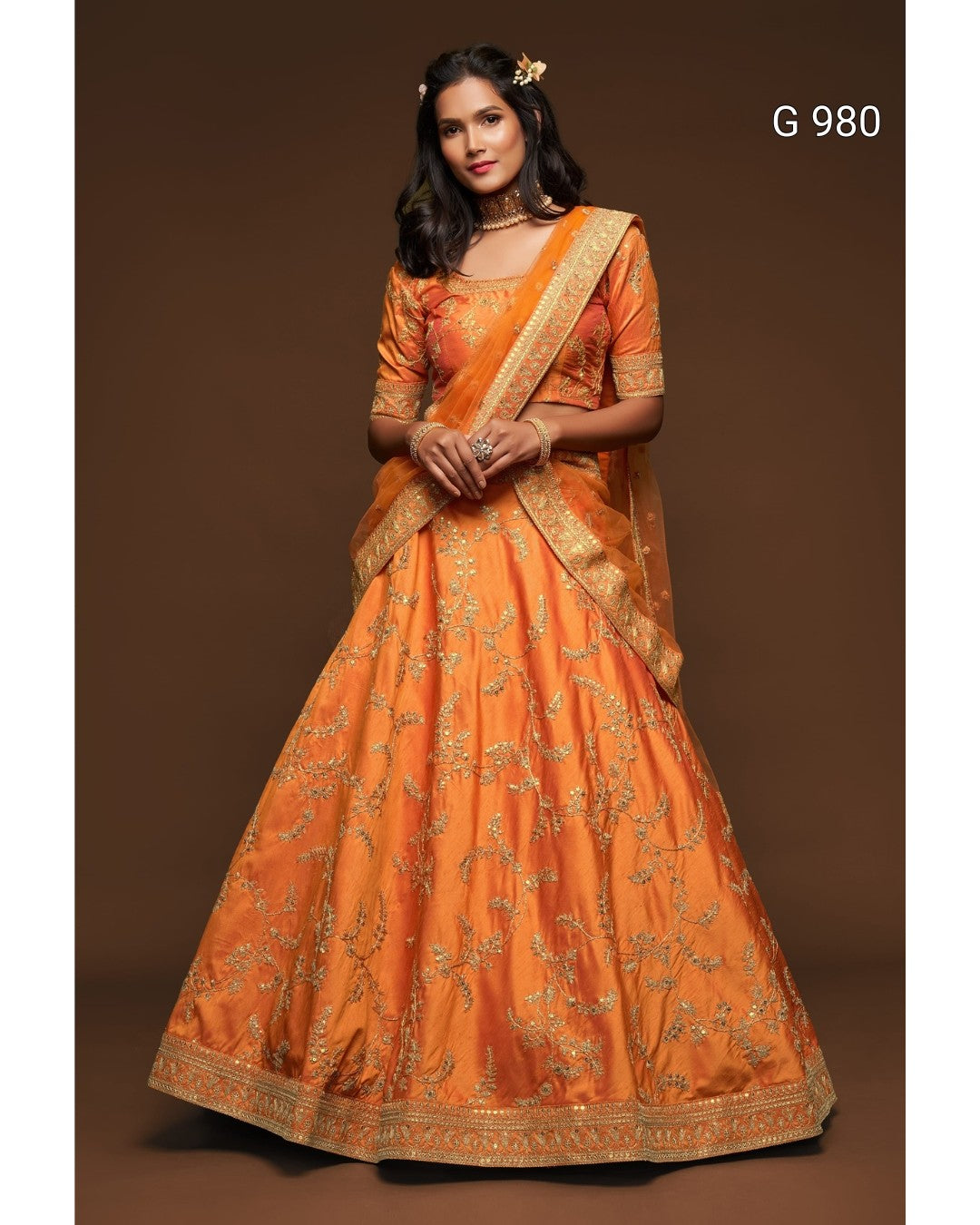 A woman models a traditional Indian orange lehenga choli set with intricate gold embroidery, featuring a flared skirt, embroidered bodice, and a matching dupatta, set against a solid brown studio background.