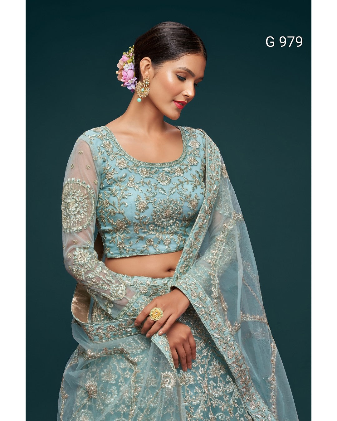 A woman modeling a light blue embroidered traditional outfit with a matching sheer dupatta, set against a dark teal background. The outfit features intricate gold embroidery on the blouse and skirt, and the model is wearing gold jewelry and a flower in her hair.