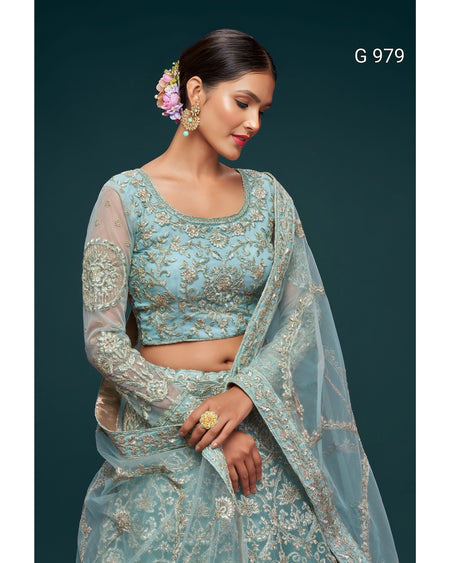 A woman modeling a light blue embroidered traditional outfit with a matching sheer dupatta, set against a dark teal background. The outfit features intricate gold embroidery on the blouse and skirt, and the model is wearing gold jewelry and a flower in her hair.