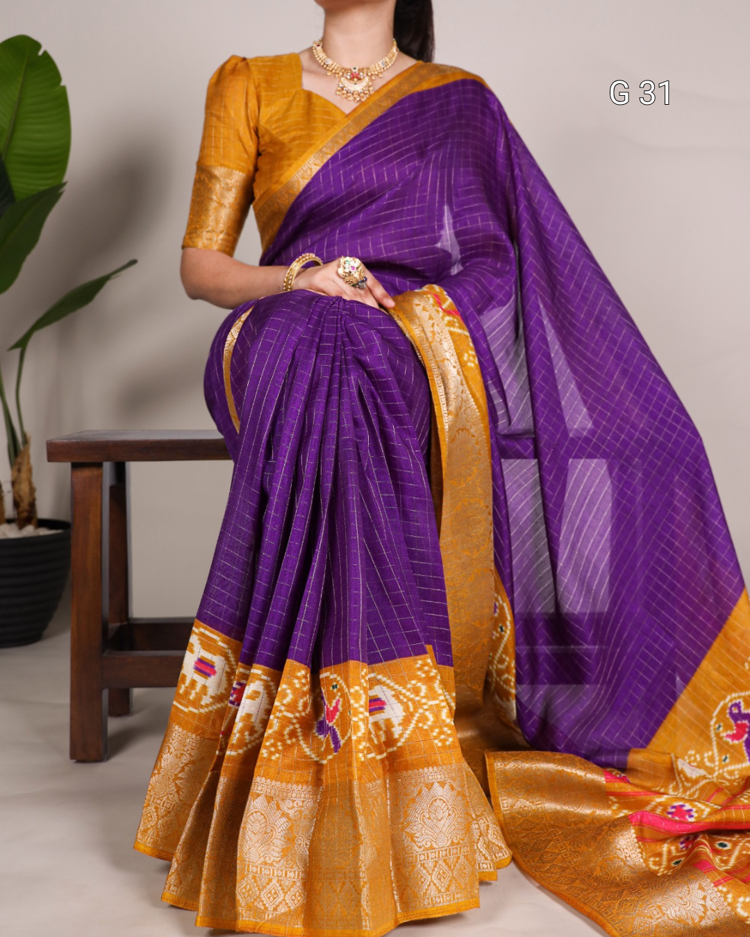 Woman wearing a purple and gold saree with a plant and chair in the background