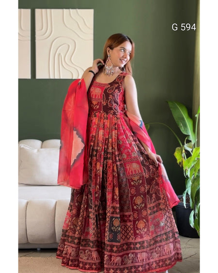 A woman models a traditional Indian salwar kameez set in a living room setting. The outfit consists of a dark red and brown printed kurta with a matching flared skirt and a bright red dupatta.