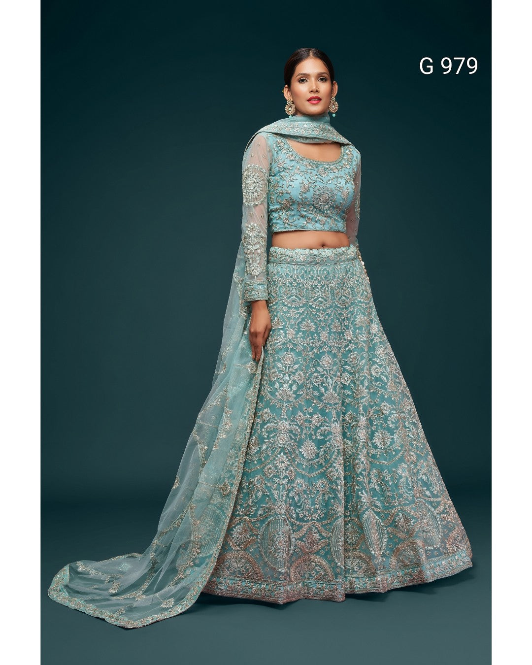 A model wearing a light blue-green embroidered traditional Indian outfit with a matching dupatta, set against a dark teal background. The outfit features a heavily embroidered crop top, a flared skirt with a wide border, and a long sheer sleeve.