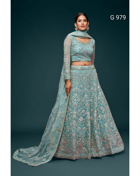 A model wearing a light blue-green embroidered traditional Indian outfit with a matching dupatta, set against a dark teal background. The outfit features a heavily embroidered crop top, a flared skirt with a wide border, and a long sheer sleeve.