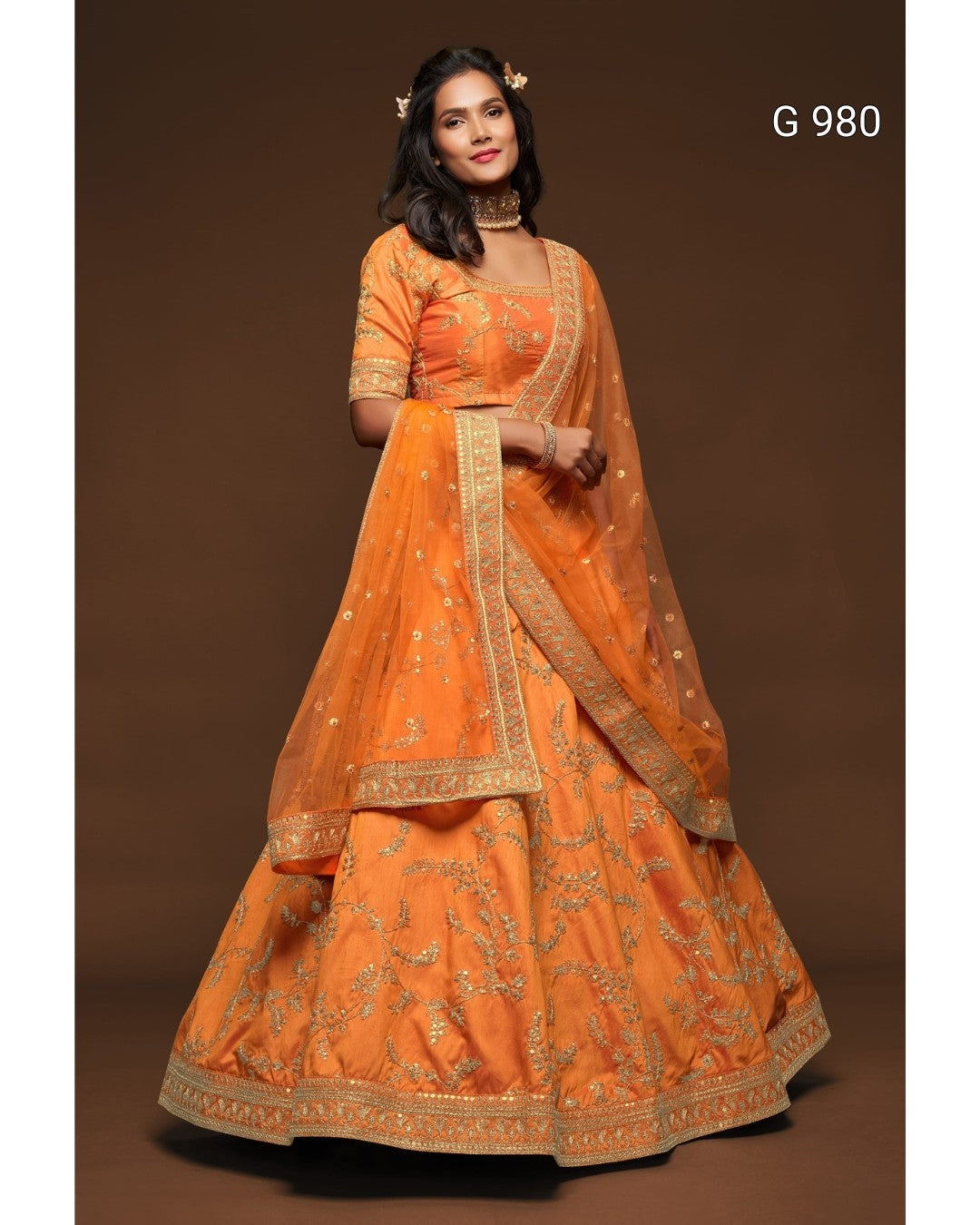 An orange traditional Indian lehenga choli dress with gold embroidery, modeled against a solid brown studio background. The text 'G 980' is visible in the top right corner.