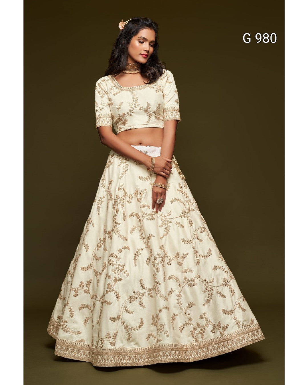A woman modeling a cream-colored traditional Indian lehenga choli set with gold floral embroidery, set against a dark olive-brown studio background. The text 'G 980' is visible in the top right corner.