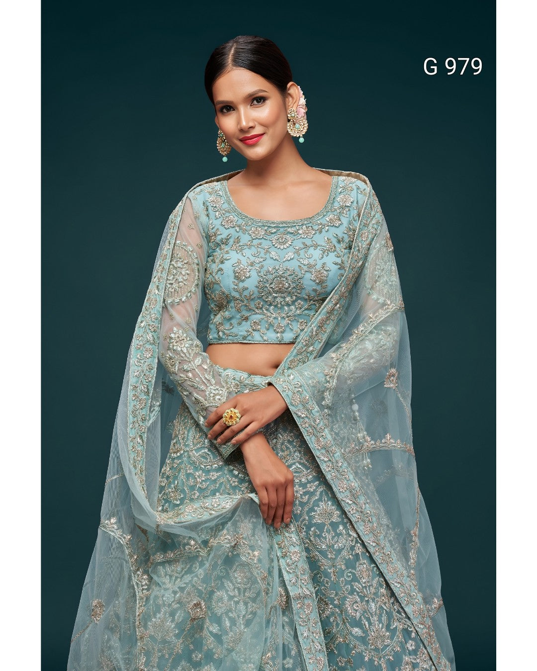 A woman models a light blue embroidered traditional outfit with a matching sheer dupatta, set against a dark teal background. The outfit features intricate gold embroidery and has the text 'G 979' printed in the top right corner.
