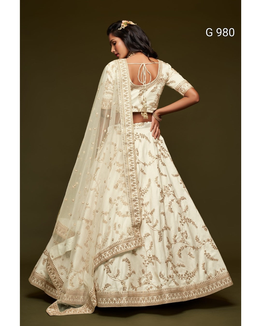 A woman modeling a cream-colored traditional Indian lehenga choli with gold floral embroidery, shown from the back against a dark brown studio background. The text 'G 980' is visible in the top right corner.