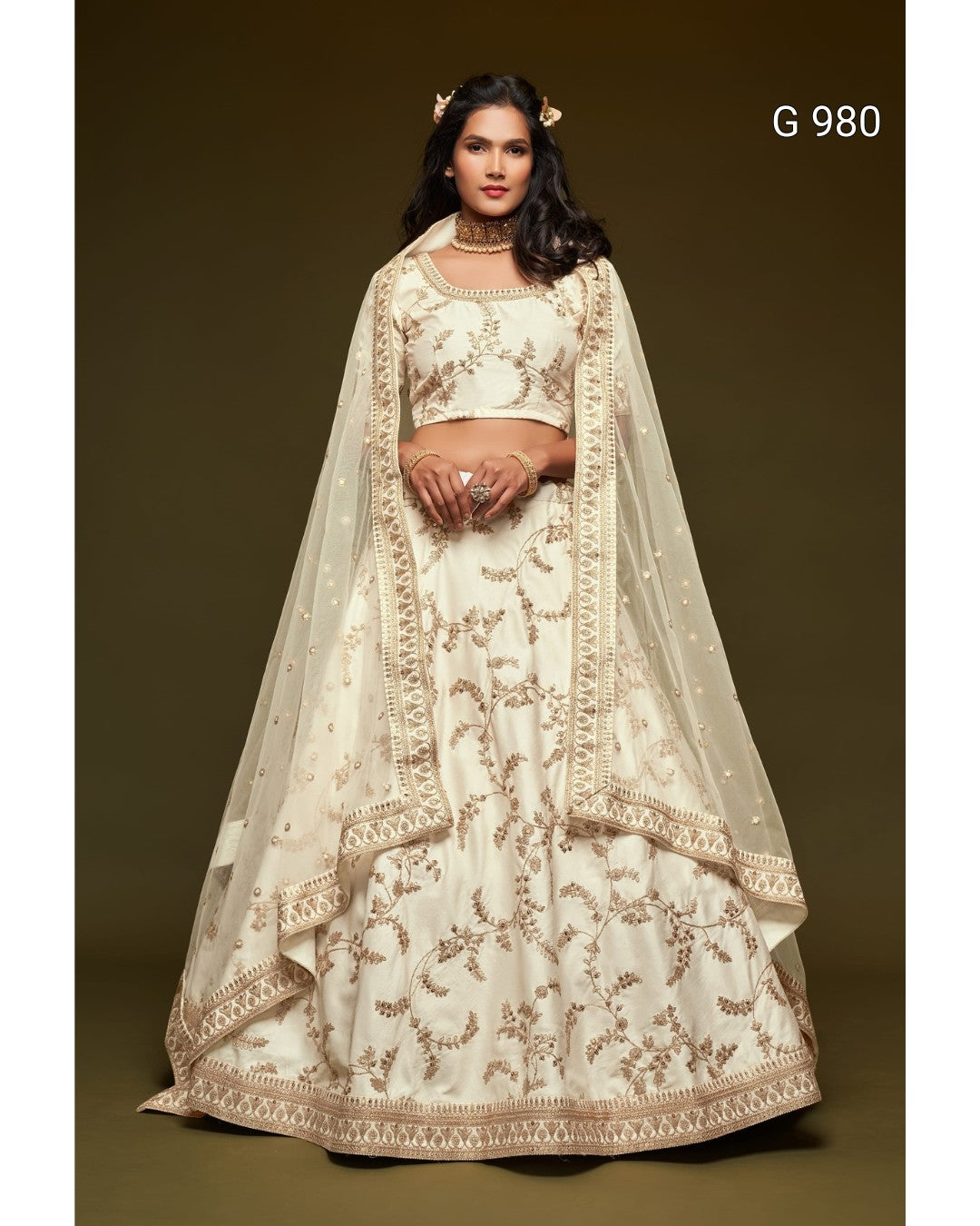 A woman modeling a traditional cream-colored lehenga choli set with intricate gold embroidery, featuring a matching sheer dupatta, set against a solid dark brown studio background. The text 'G 980' is visible in the top right corner.
