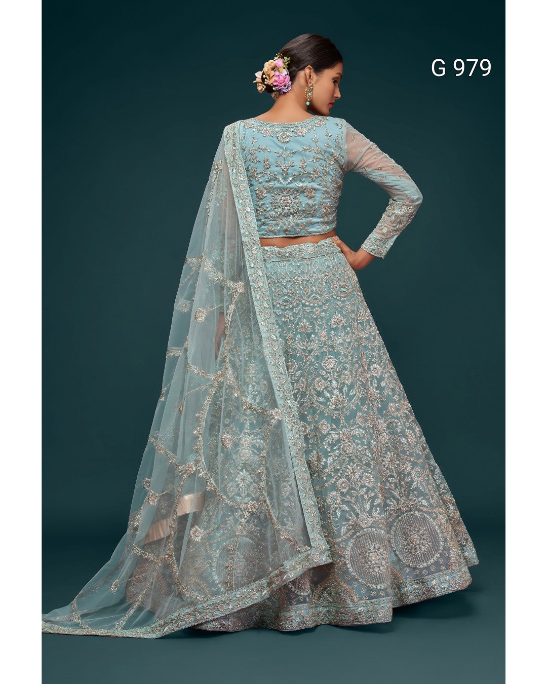 A woman modeling a light blue embroidered lehenga choli set with a matching dupatta, shown from the back against a dark teal studio background. The outfit features intricate silver and gold floral embroidery, and the model has her hair styled with a pink flower accessory.