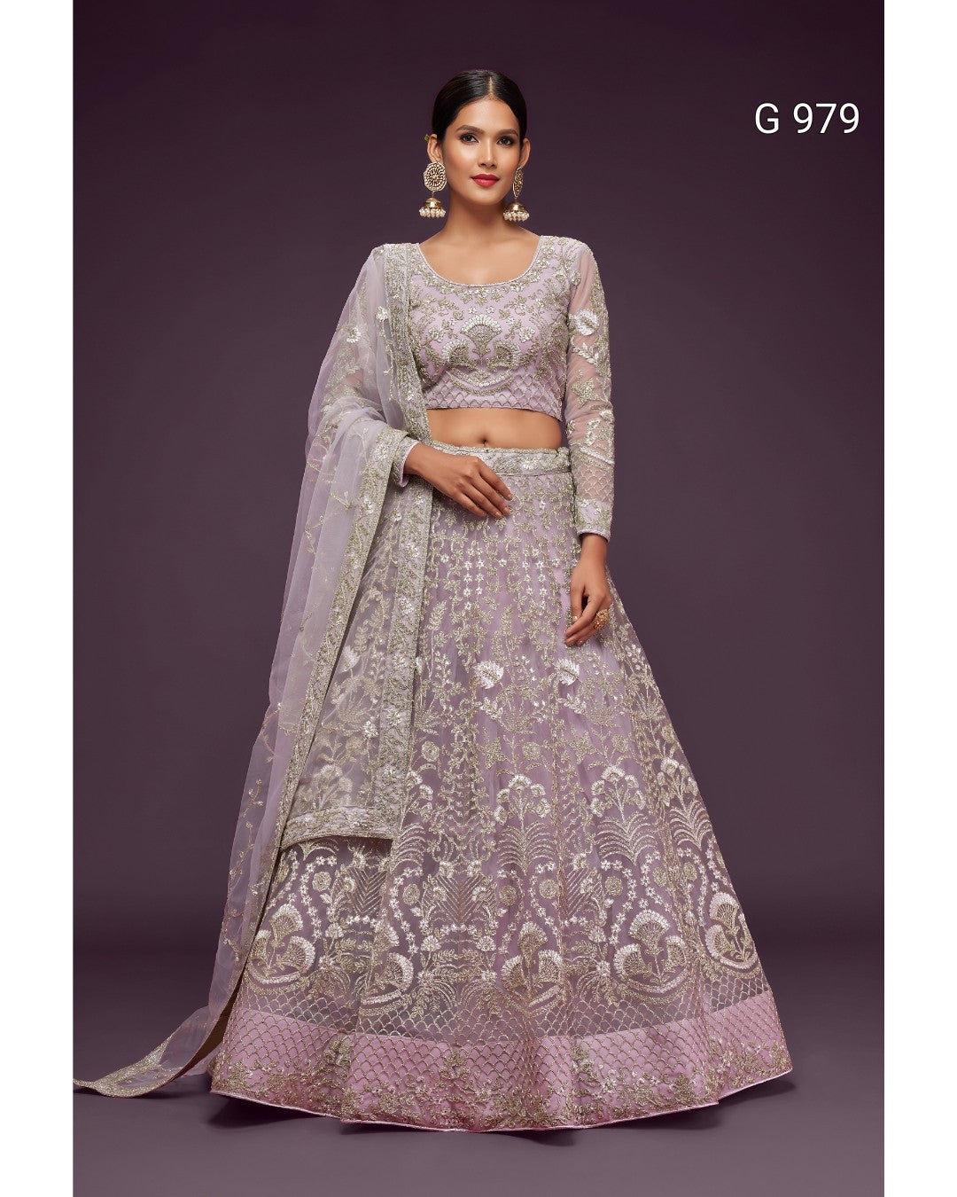 A woman modeling a lavender and silver embroidered Indian ethnic outfit, featuring a flared lehenga skirt, a matching embroidered crop blouse, and a sheer dupatta, set against a solid dark purple background.