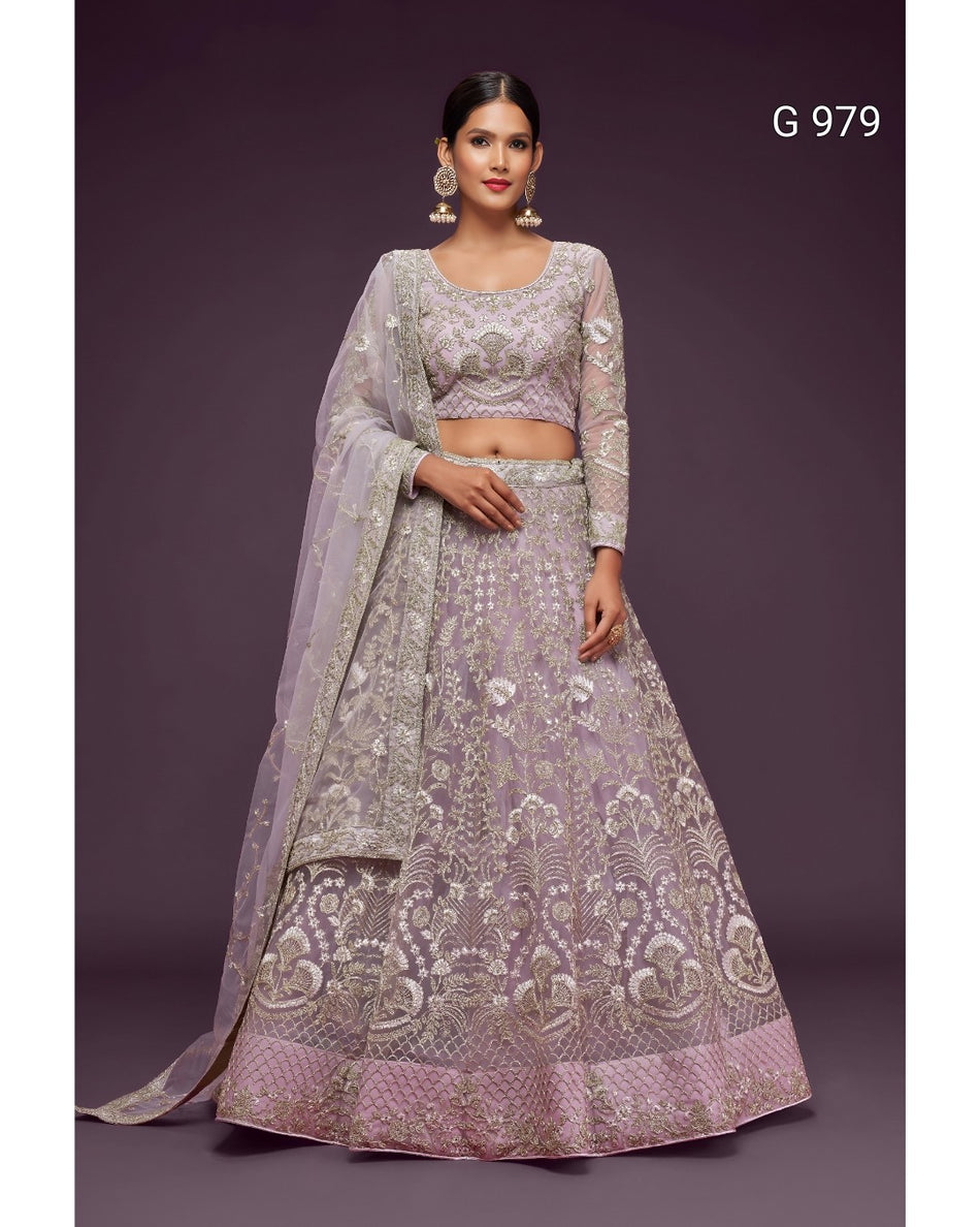 A woman modeling a lavender and silver embroidered Indian ethnic outfit, featuring a flared lehenga skirt, a matching embroidered crop blouse, and a sheer dupatta, set against a solid dark purple background.