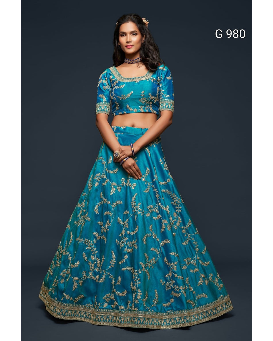 A woman modeling a teal blue lehenga choli set with intricate gold floral embroidery, set against a dark gray studio background. The text 'G 980' is visible in the top right corner.