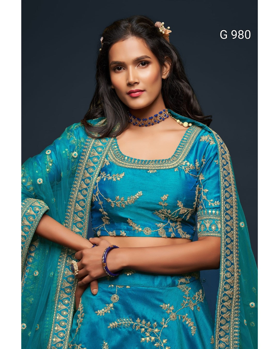 A woman modeling a teal blue ethnic outfit with gold floral embroidery, featuring a matching blouse, skirt, and dupatta, set against a dark studio background with the text 'G 980' in the top right.