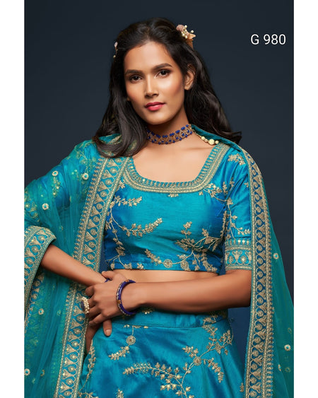 A woman modeling a teal blue ethnic outfit with gold floral embroidery, featuring a matching blouse, skirt, and dupatta, set against a dark studio background with the text 'G 980' in the top right.