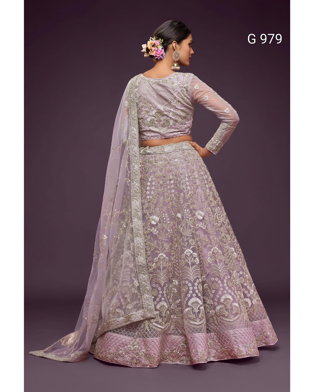 A woman modeling a lavender lehenga choli set with intricate silver embroidery, shown from the back against a dark purple studio background. The set includes a heavily embroidered skirt, a matching blouse, and a sheer dupatta.