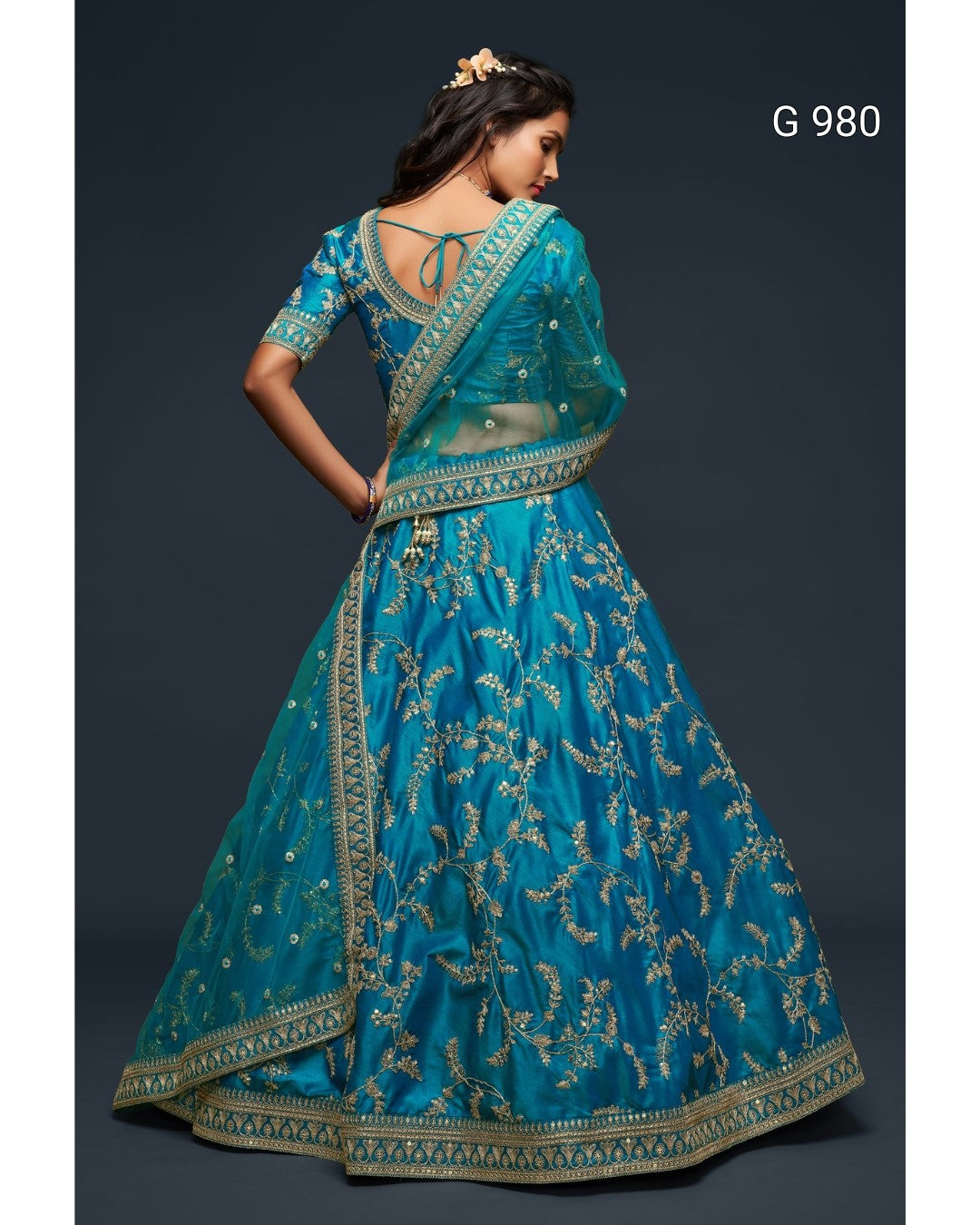 A woman modeling a teal blue lehenga choli with intricate gold embroidery, shown from the back against a dark gray studio background. The text 'G 980' is visible in the top right corner.